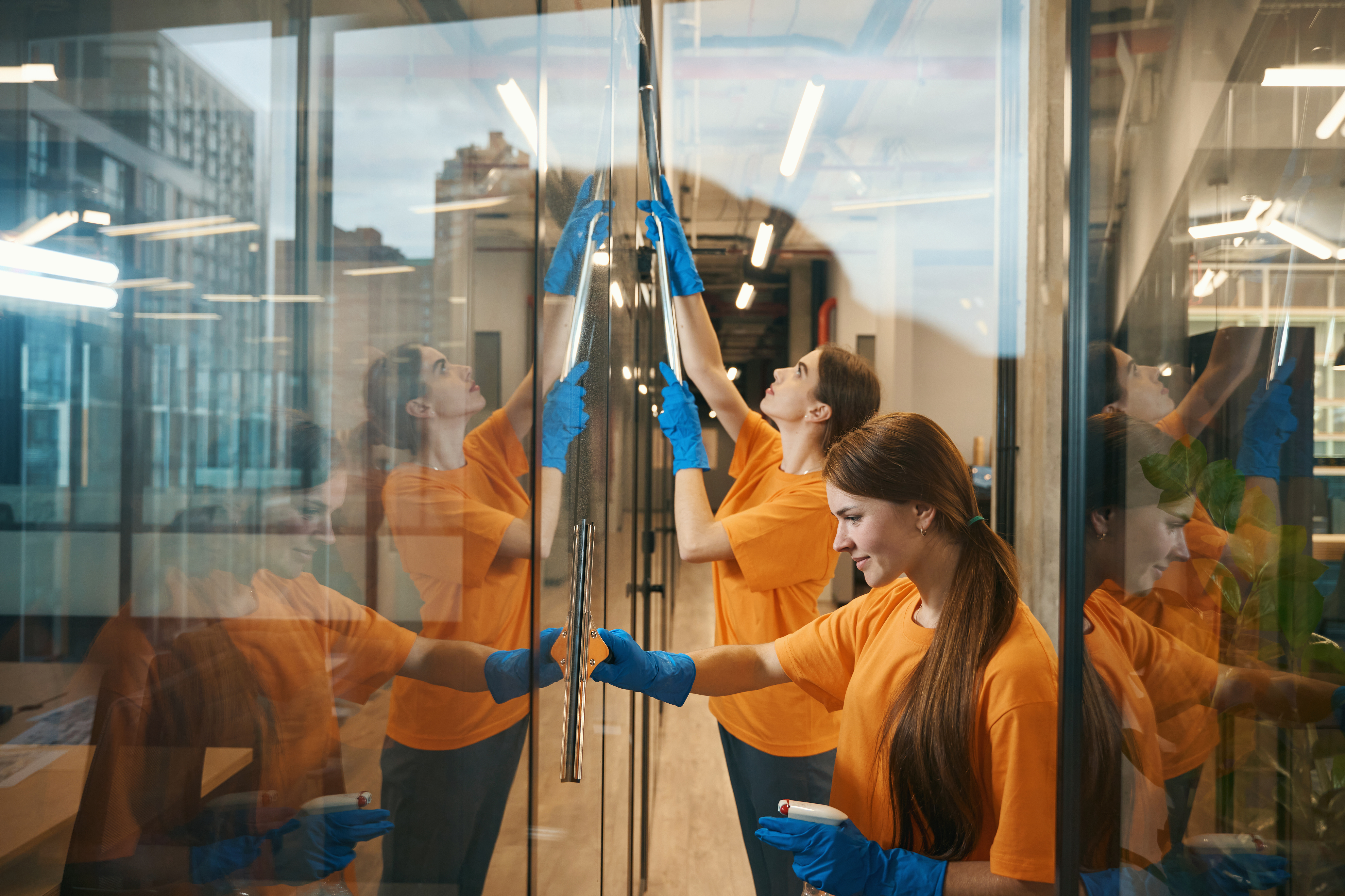 Young cleaners clean mirrored windows coworking spacer, they in simple uniforms